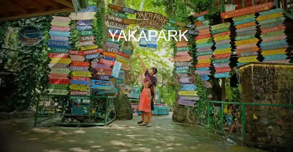 Birçok yer için cennetten bir köşe deyimi kullanılır ki gerçekten de ülkenin çoğu yeri cennetin yeryüzündeki yansıması gibi. Ancak tasvir ve anlatılanlara baktığımızda cennet sanki Yakapark’ın tanımı gibi. İster Fethiye’de tatil yapıyor olun isterseniz Kaş ya da Kalkan’da, hepsine o kadar yakın ortak bir lokasyondakiYakapark, ziyaret etmeden tatilinizi sonlandırmak istemeyecek, görmediyseniz de hayıflanarak tekrar tatil planı yapmaya başlayacaksınız. Yeşilin tüm renklerini içinde barındıran bir doğa düşünün tam ortasında serin ve sakinleştiren sesiyle şelale aksın gölleştirdiği sularıyla. Kulağa çok güzel geldiğinin farkındayız, bu tecrübeye dahil olmak istemez misiniz? Doğayı seviyorsunuz Yakapark’tan ayrılmak çok zor gelecek sizlere. Farklı türdeki kuş cıvıltılarının şenlendirdiği bu bölge, ruhun dinlenip arındığı saklı kalmış cennetlerden biri. Devasa ağaçların gölgesinde biraz mola verip dinlenebilir, molanıza eşlik eden huzur ile de tüm yorgunluğunuzu unutacaksınız. Küçük bir mola diye uğrayacağınız Yakapark, sizi ağırlamak için çok bir çaba sarf etmeyecek. Daha ilk dakikalardan itibaren sizi kuşatmaya başlayan oksijenin oluşturacağı etkiye kendinizi teslim etmeye hazır olun. Usul usul ancak davetkar hamaklar, manzaranın tadını iyice çıkarasınız diye taraça şeklindeki seyir basamakları, yoğunluğuyla çağlayan su kanalları ve havuza bürünmüş gölleriyle gözünüzü kapatın sesi ve huzurun dinleyin. Tatiliniz anlam kazanmaya başladı bile. Ege kıyılarının en güzel özelliklerinden biri o sıcakkanlı kadınlarının hazırlamış olduğu birbirinden nefis saçta pişen gözlemelerin her durakta rastlıyor olmak. Yakapark’ta, birbirinden renkle sedirlerin oluşturmuş olduğu salaş ve doğal mekanlarda oturup bir şeyler atıştırıp dinlenmeniz de mümkün. Ancak buraya gelmişken gözlemelerin tadına bakmadan ayrılmayın. Ege ve gözlemeleri, tatil anılarınıza bir madde daha eklemenize neden olacak. Çoğu yol üstünde karşılaşacağınız gözlemeler için oluşturulmuş gözleme mekanları da mevcut. Bölgede Alabalık da revaçta. Kanal sularında yaşayan ve doğal yöntemlerle beslenen Alabalıkların da tadı doğal olarak damağınızdan kolay silinmeyecek bir lezzet. Göl ve kanalda yüzen balıkları izlerken, balık atıştırmak isteyebilirsiniz. Yakapark başlı başına bir destinasyon ancak buraya gelmişken yakınlarındaki diğer gezilecek yerleri de ziyaret etmeniz mümkün. Bunların başında, Kaunos, Pınara, Letoon, Xantos, Patara plajı ve antik kentler, kaya mezarları, Fethiye tiyatrosu ve müzesi, Kayaköy, Ölüdeniz Milli Parkı, Katrancı Koyu, Kızlar Koyu, Göcek ve Kaş’ı sayabiliriz. Gündüz uzayıp giden bembeyaz kumlarla örtülü plajlarda denizin ve güneşin tadını çıkarırken, akşamında doğal güzellikler sunan tabiatın eşsiz manzarası eşliğinde dinlenebilirsiniz. Tatilinizde her türlü aktivitelerin olması hem tatilinizi daha canlı kılacak hem de birçok alternatifle tatilinizi değerlendirmiş olacaksınız. Zaten Kaş, Kalkan ya da Fethiye’de tatildeyseniz Yakapark’a uğramadan dönmeyeceksiniz. Yakapark’ın girişinde rastlayacağınız renkli ve küçük eğlenceli yön tabelaları daha ilk dakikadan ne kadar güzel bir yere geldiğinizi kanıtlar gibi.